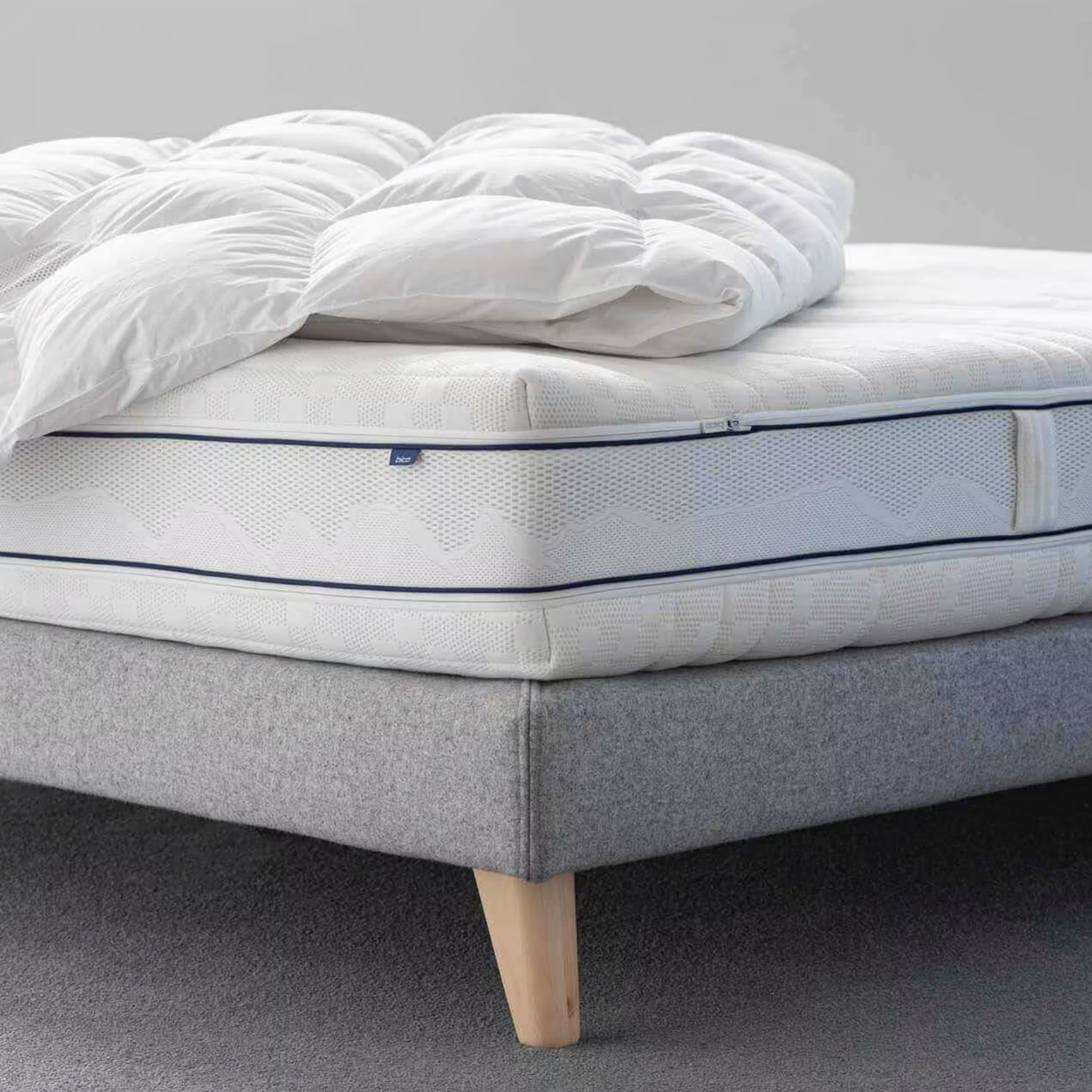 Weiße Matratze Clima Luxe mit feiner Linienstruktur, seitlicher grauer Griff und blauer Biese, liegt auf grauem Bettgestell.